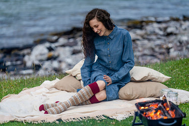 A woman sits on a cream blanket on grass by the water, wearing a blue denim dress and patterned knee-high socks. A small fire pit with glowing embers sits nearby, with rocks and water in the background.