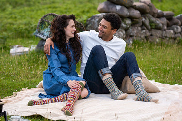A man and a woman sitting on a cream blanket on grass, smiling at each other. The woman wears a blue shirt and colorful fairisle knee-high socks, and the man wears a white t-shirt, dark jeans, and striped socks.