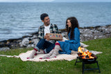 A man and a woman sit on a blanket by the ocean, sharing a cardboard gift box with colorful socks. A small fire burns in a black metal campfire bowl on the grass beside them, with rocks and water in the background.