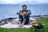A man sits on a blanket by the ocean with a small fire burning in a black grill. He is wearing a green and black plaid flannel shirt, blue jeans, and patterned knit socks.