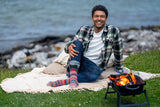 A man sitting on a blanket by the water, wearing a plaid shirt, jeans, and colorful striped socks, with a small portable fire pit on the grass in front of him.
