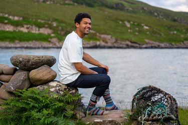 A man sitting on a rock by the water wearing a white t-shirt, dark jeans, and colorful fairisle socks, with a lobster trap on the ground nearby and a green hill in the background.