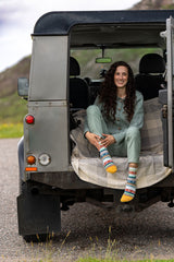 A woman sits in the open back of a gray off-road vehicle on a gravel path, wearing a sage green matching lounge set and colorful striped socks.