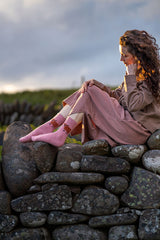 A woman sits on a stone wall outdoors, wearing a dusty pink outfit and pink socks with a brown cow print. The background shows a cloudy sky and green grassy hills.