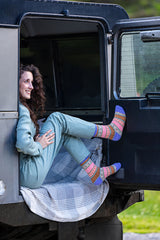A woman with curly brown hair sits in the open doorway of a dark vehicle, wearing a light blue sweatshirt and matching sweatpants with colorful patterned socks. A plaid blanket is draped over the step.