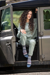 A woman with long curly brown hair sits in the open doorway of a dark vehicle, wearing a sage green matching lounge set and fairisle patterned socks.