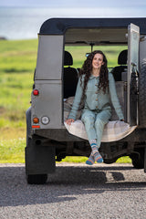 A woman with long curly brown hair sits on the open rear of a gray off-road vehicle on a gravel road, wearing a matching sage green two-piece outfit with a plaid blanket and colorful socks, with a grassy field in the background.