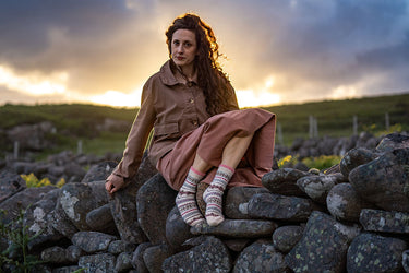 A woman with long curly brown hair sits on a stone wall outdoors, wearing a brown jacket and dusty pink skirt with patterned socks, against a dramatic cloudy sky at sunset.