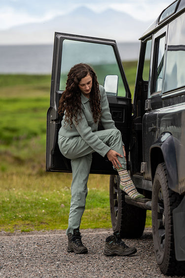 A woman in a matching sage green tracksuit stands by the open door of a black off-road vehicle, adjusting a striped knit sock. The background shows a blurred green landscape and distant mountains.